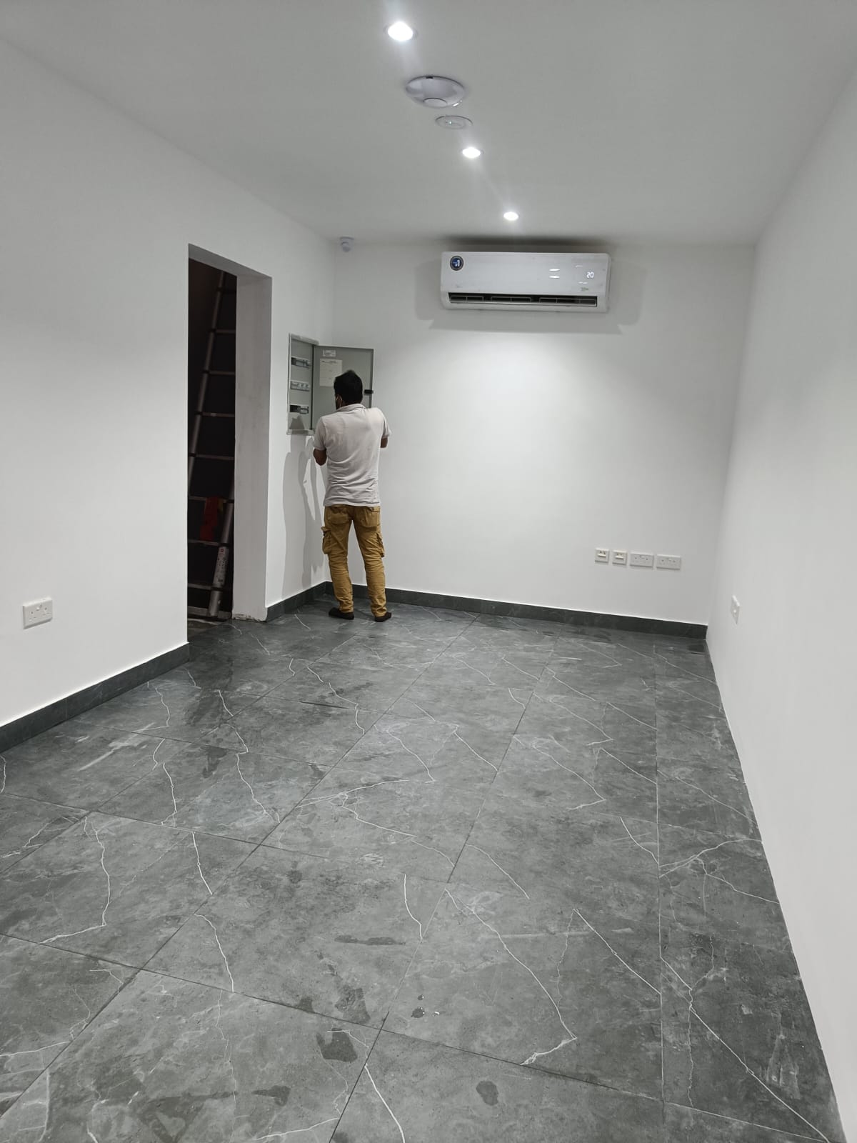Technician at an open electrical panel in a renovated office with marble-effect floor and recessed ceiling lights